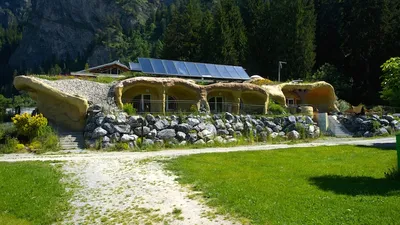 Das Erdhaus von aussen: gerundete, verputzte Wände mit Grasdach, Sonnenkollektoren darauf, davor eine Mauer aus Findlingen